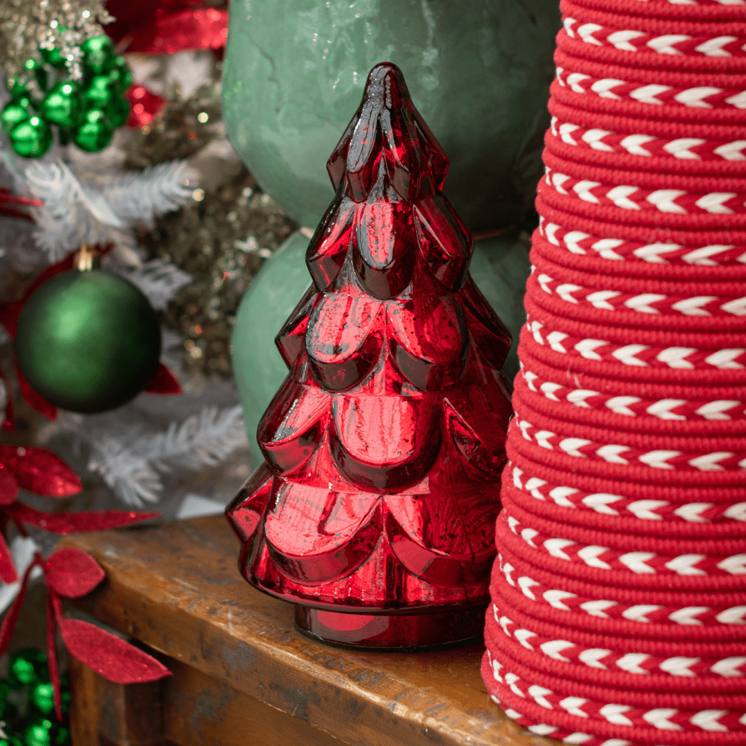 A red glass decorative Christmas tree sits on a wooden table with a decorated white, green, and red artificial Christmas tree behind it.
