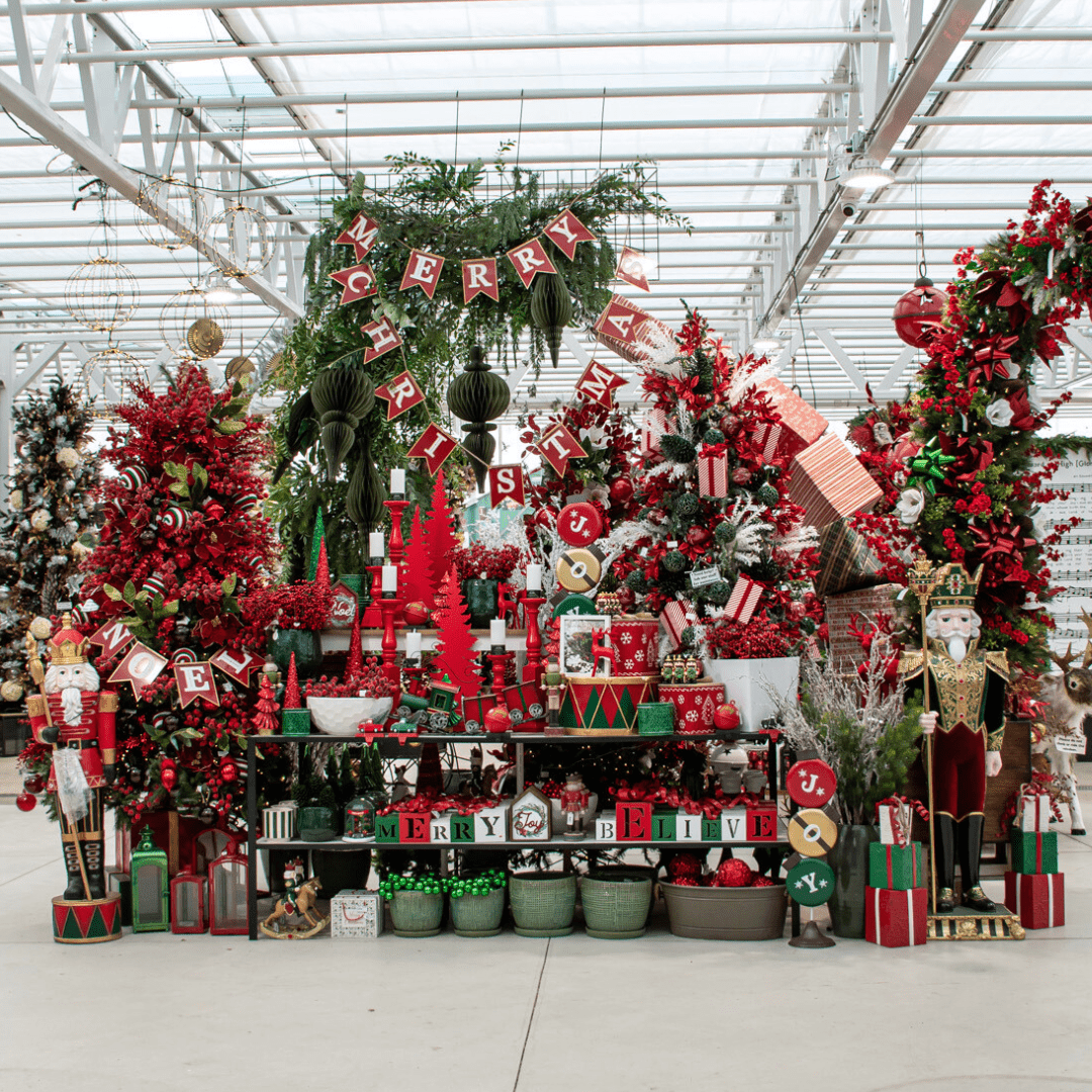 A traditional red, green, and white Christmas display in a greenhouse with multiple artificial decorated Christmas trees and a large metal sign that says 'Merry Christmas'.