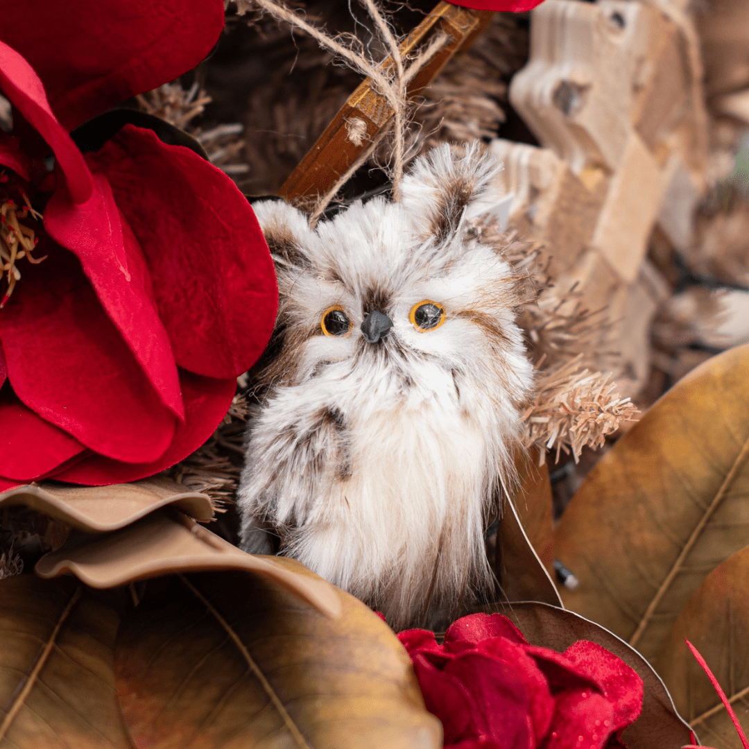 A fuzzy white and brown owl ornament hangs in a beige artificial Christmas tree with other brown and red accents.