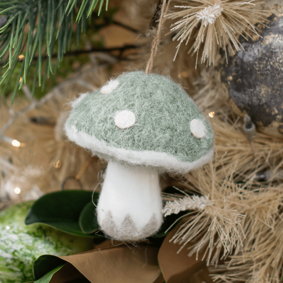 A felt white and sage mushroom ornaments hangs from a beige artificial Christmas tree.