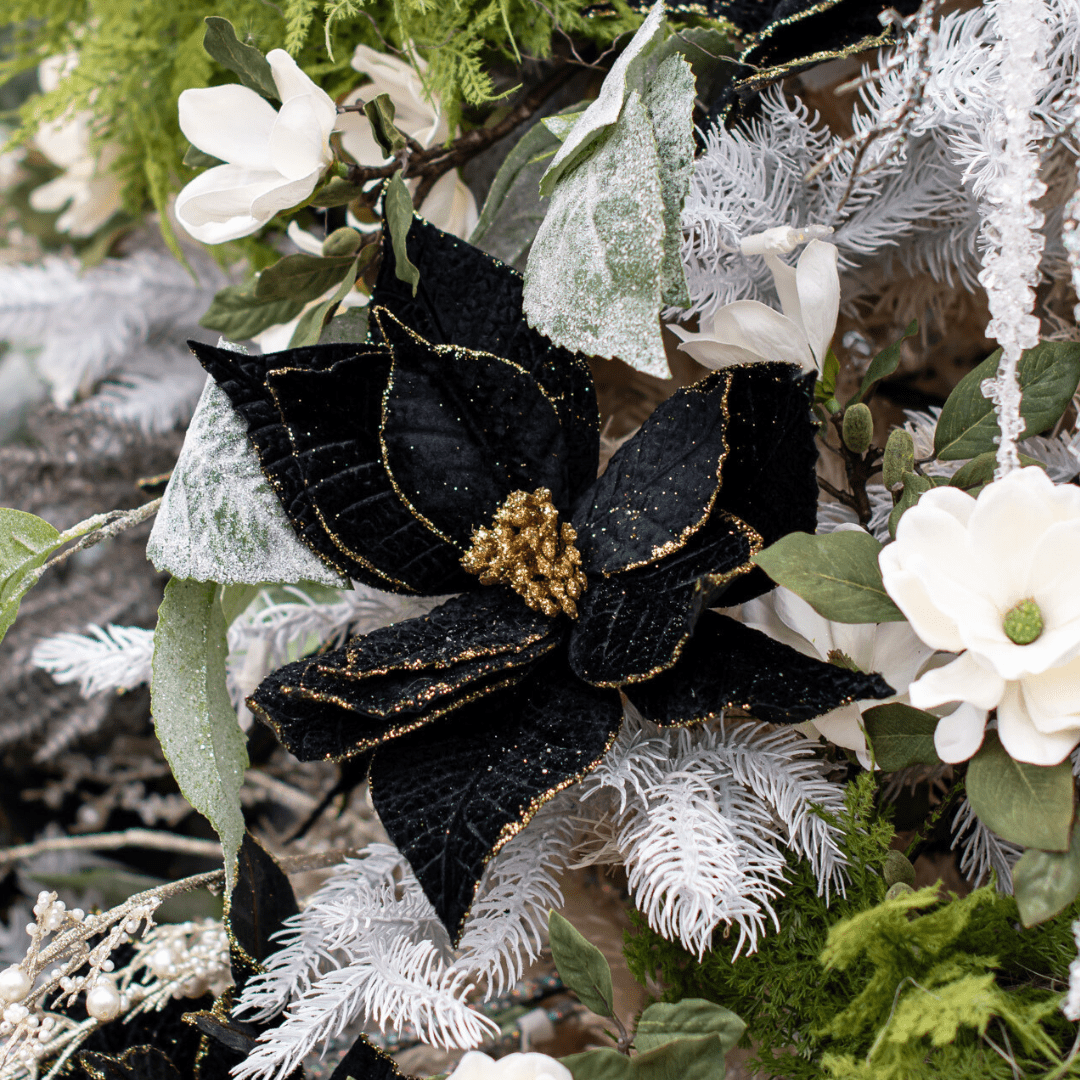 A faux black and gold velvety poinsettia pick in an artificial white Christmas tree with other artificial greenery around it.
