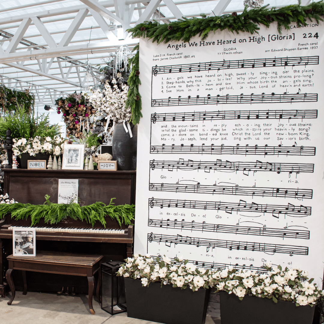 A black and white Christmas display in a greenhouse with a large painted sheet music of 'Angels We have Heard on High' beside a dark wood piano.
