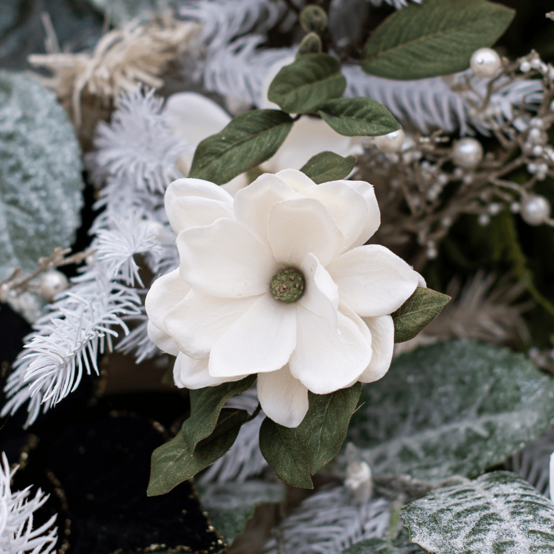 A faux white magnolia flower pick in a white artificial Christmas tree with frosted-looking green leaves around it.