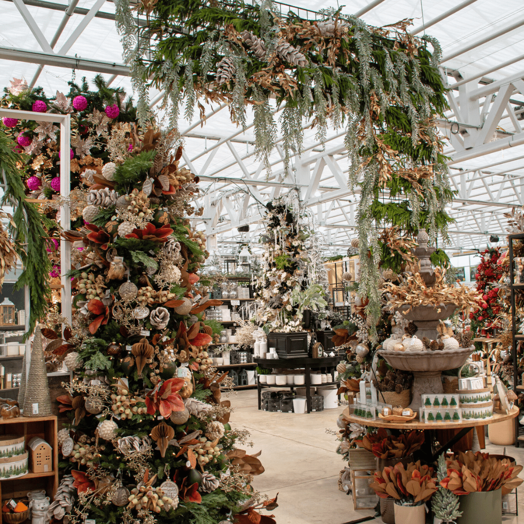 A green, brown, beige, and natural-coloured Christmas display in a greenhouse with artificial Christmas trees, a tiered resin fountain, an L-shaped greenery arch, etc.