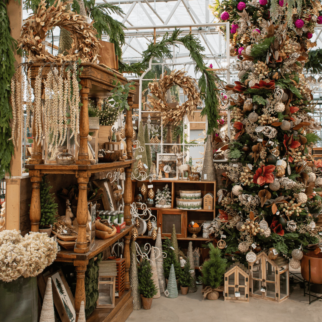 A green, brown, beige, and natural-coloured Christmas display in a greenhouse with artificial Christmas trees, a large white-framed mirror, and a wooden shelf with decor.