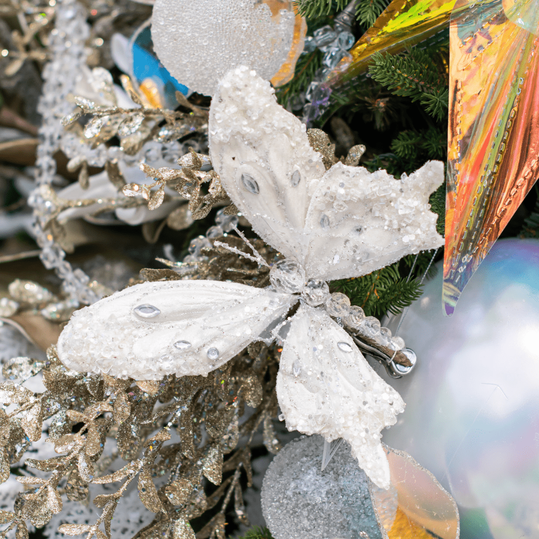 A white, sparkly butterfly pick in an artificial green Christmas tree surrounded by other white and iridescent decorations.