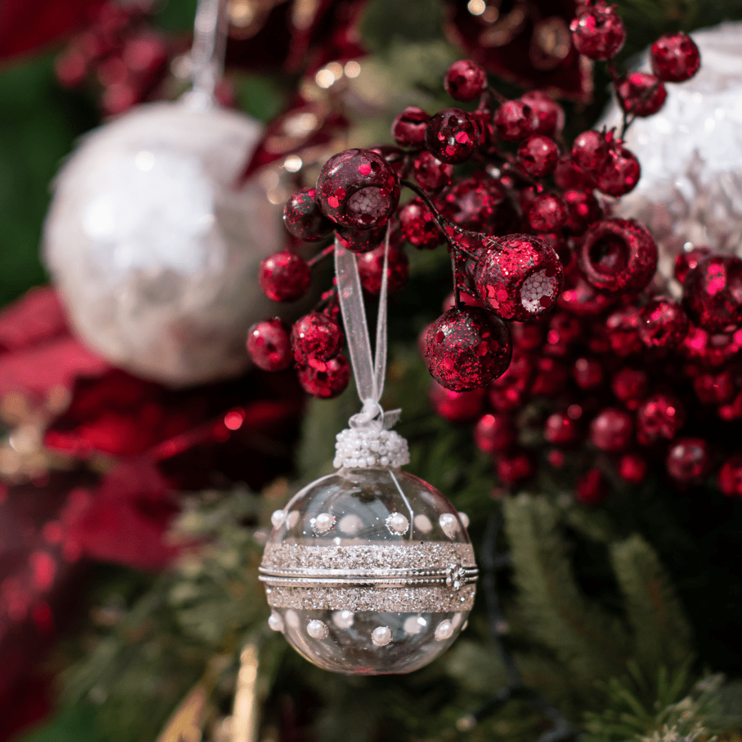 A small round clear and sparkly plastic ornament hangs from a burgundy berry pick in an artificial green Christmas tree.