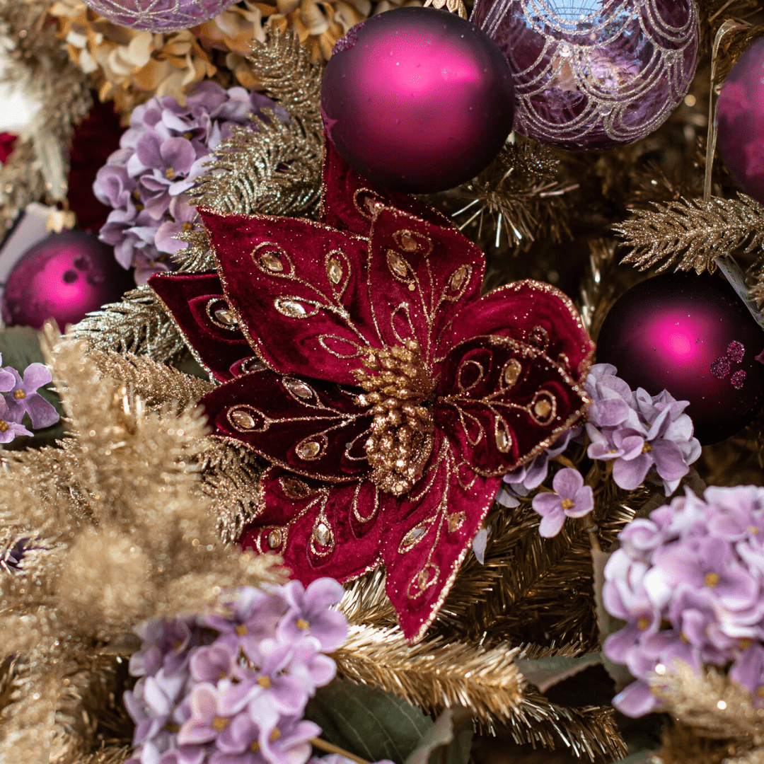 A dark red and gold sparkly poinsettia pick in a gold sparkly artificial Christmas tree with other purple and berry-coloured decorations around it.
