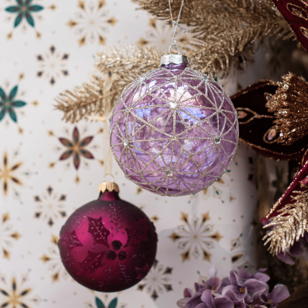 Two round ornaments, one light purple and one dark berry-coloured, hang from a gold sparkly artificial Christmas tree.