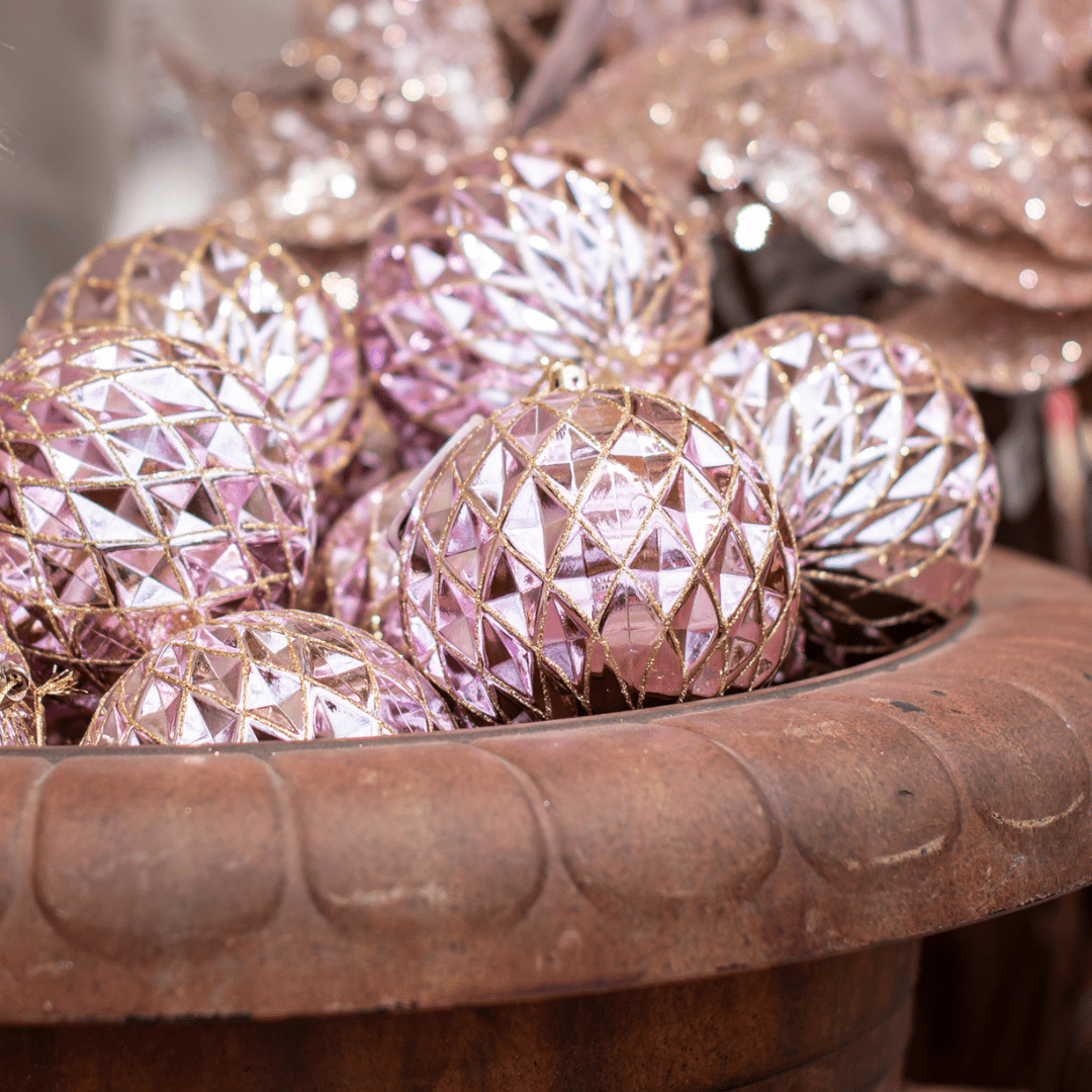 Light pink and gold round ornaments with a diamond pattern are displayed on top of one another in a rustic urn.