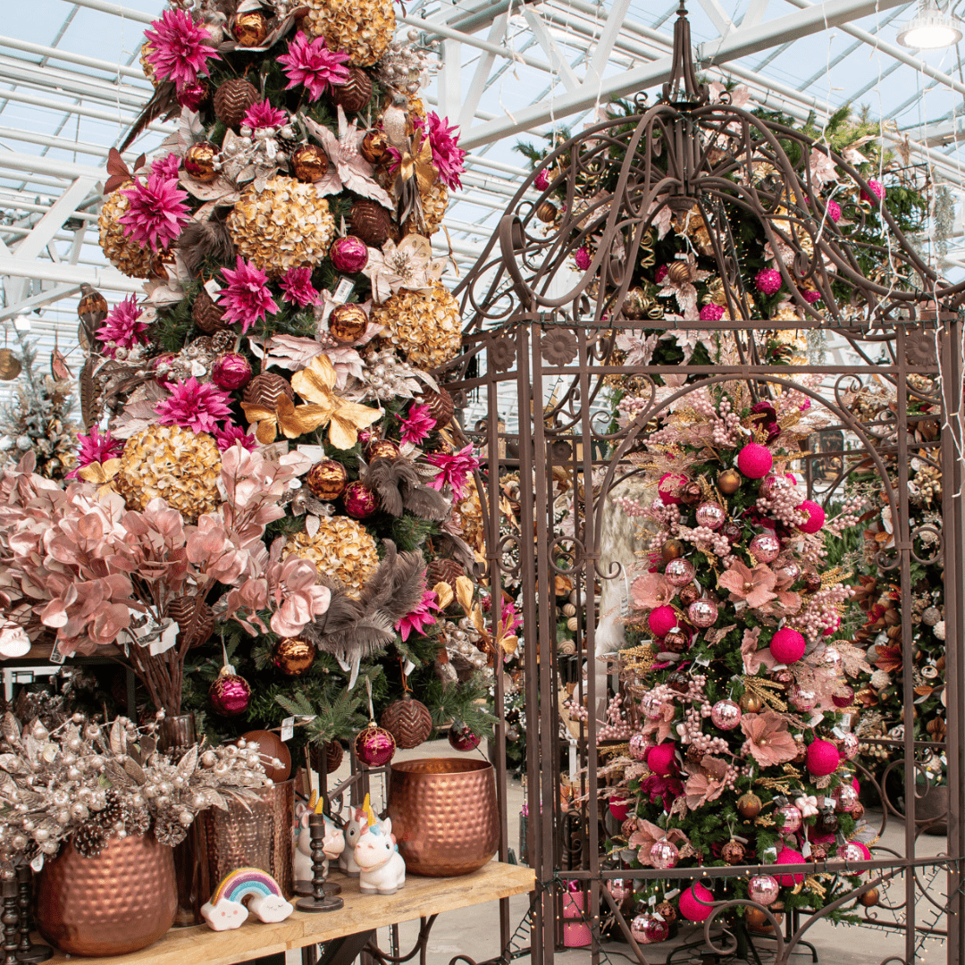 A pink, brown, and gold Christmas display in a greenhouse with a large fake furry reindeer, an upside down artificial Christmas tree, a brown metal gazebo, etc.