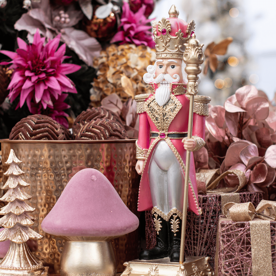 A pink and gold nutcracker stands on a tabletop with other pink, gold, and brown holiday decor.