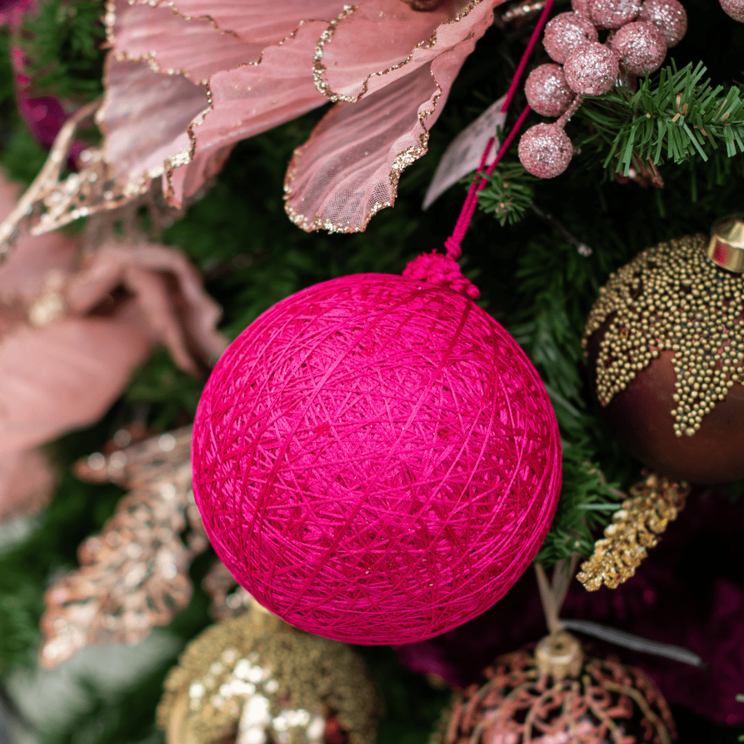 A bright pink yarn ball ornament hangs in an artificial green Christmas tree with other pink and brown decorations around it.
