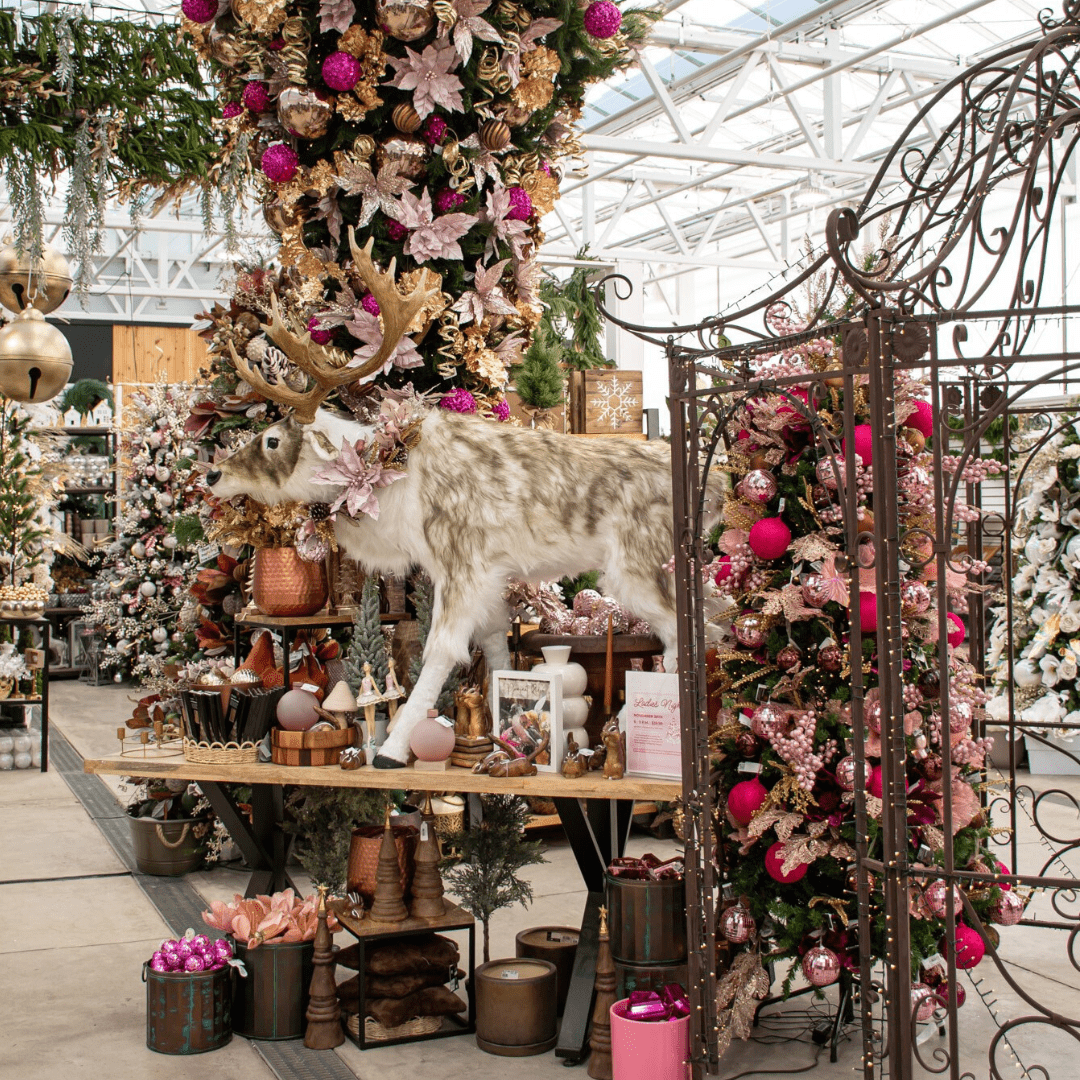A pink, brown, and gold Christmas display in a greenhouse with a large fake furry reindeer, an upside down artificial Christmas tree, a brown metal gazebo, etc.