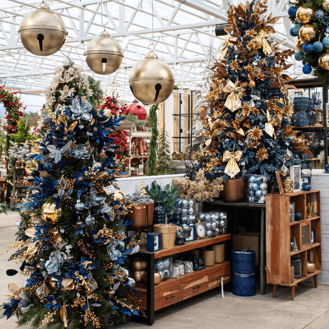 A blue, gold, and bronze Christmas display in a greenhouse with artificial Christmas trees, metal hanging bells, boxed ornament sets, etc.