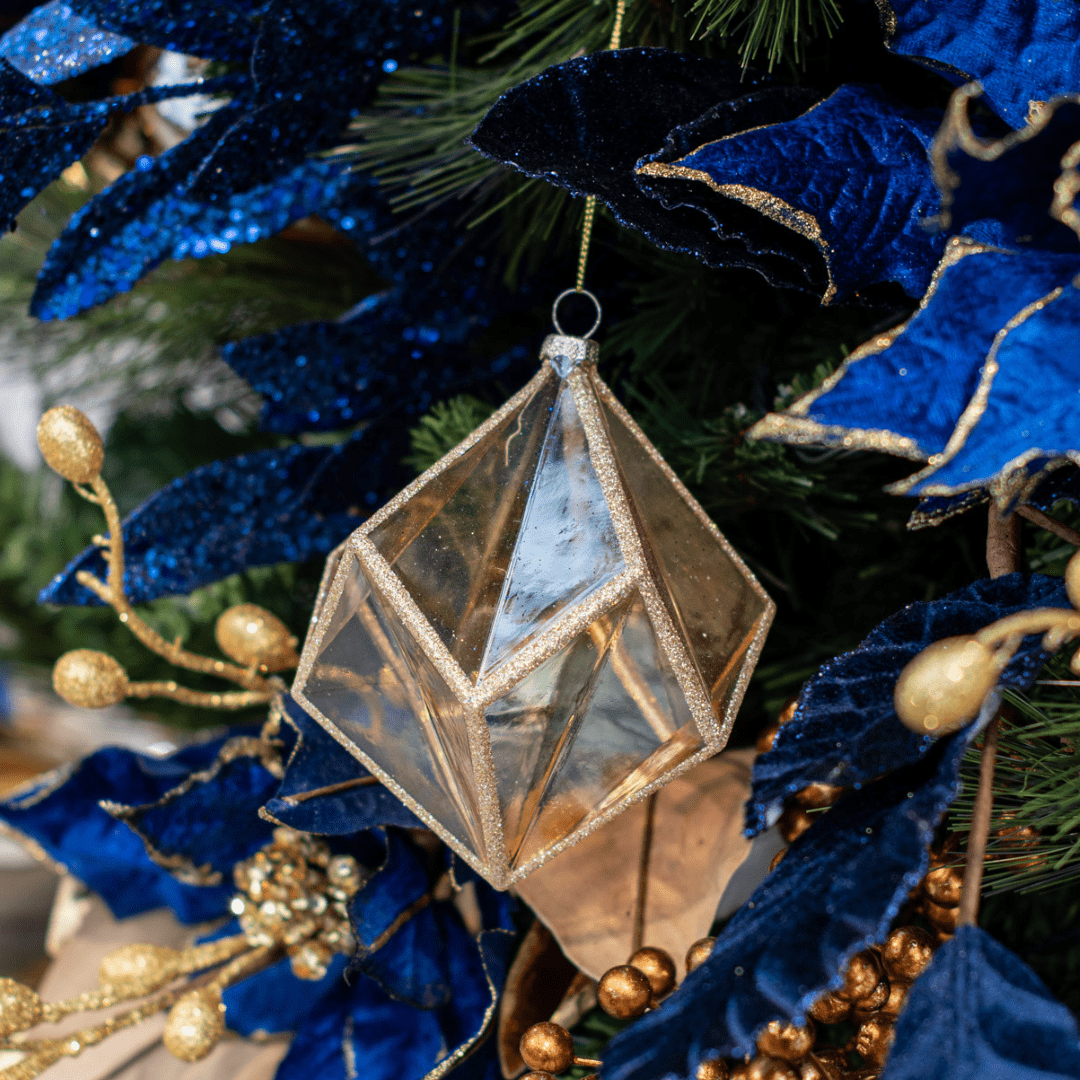 A gold geometric ornament hangs in an artificial green Christmas tree with other blue and gold decorations around it.