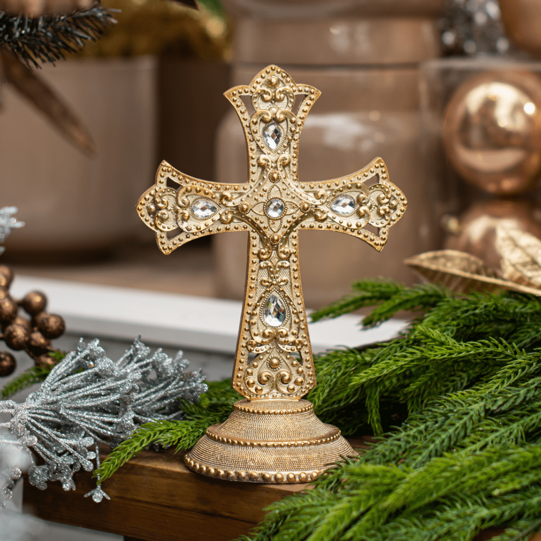 A gold table-top cross with faux diamond accents sits on a wooden mantel with artificial greenery behind it.