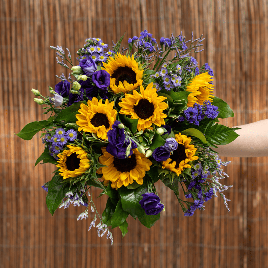 An outstretched hand holds a yellow and purple fresh floral bouquet with sunflowers.