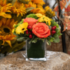 An orange, yellow, red, and lime green floral arrangement in a glass cube sits on a bale of straw.