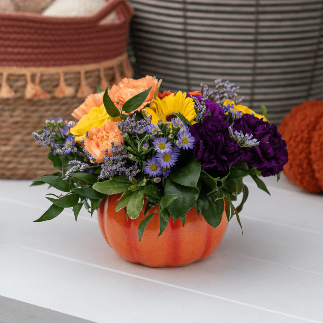 A yellow, peach, and purple fresh floral arrangement in an orange ceramic pumpkin-shaped pot sits on a white wooden table.