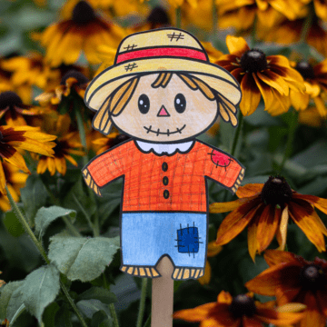 A paper scarecrow on a popsicle stick coloured with orange, blue, and yellow pencil crayons is displayed in front of fall flowers. 