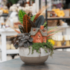 A fall-themed indoor mixed planter in a brown and cream ombre pot with a colourful croton, dark purple-black peperomia, trailing green baby tears, as well as decorative light brown wheat stems and an orange and brown brick-style ceramic house. It sits on a dark brown wooden table.