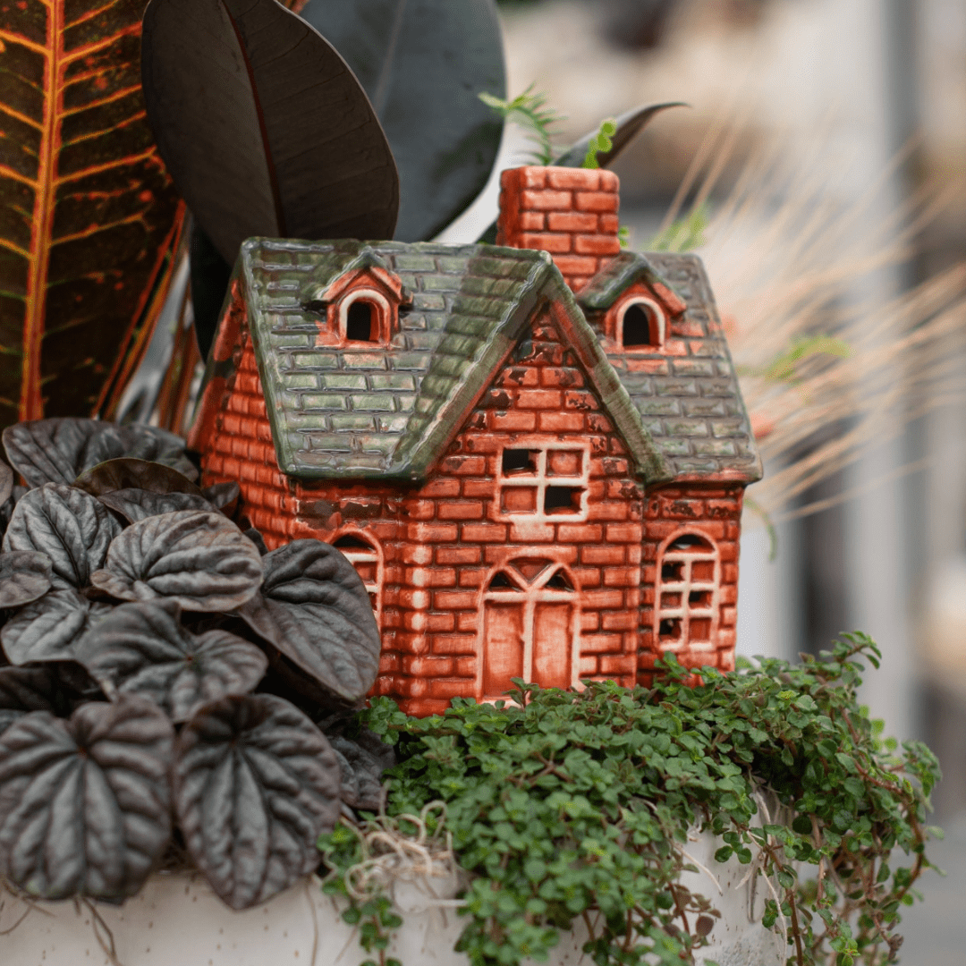 A close-up of an orange and brown brick-style ceramic house in a fall-themed indoor mixed planter.