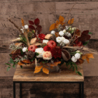 A fall floral centerpiece with rich, earthy tones, including dahlias, mums, hypericum berries, roses, lisianthus, foraged-looking branches, pods, wheat stems, and artificial apples. It sits on a wooden table in front of a faux wood backdrop.