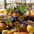 An outstretched hand holds a floral bouquet with peach, copper, and rust-coloured spider mums, amaranthus, carnations, roses, along with blue delphinium and thistle in front of a pumpkin display.