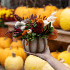 An outstretched hand holds a neutral brown, white, and burgundy fall floral arrangement in a cream and brown striped ceramic pumpkin.