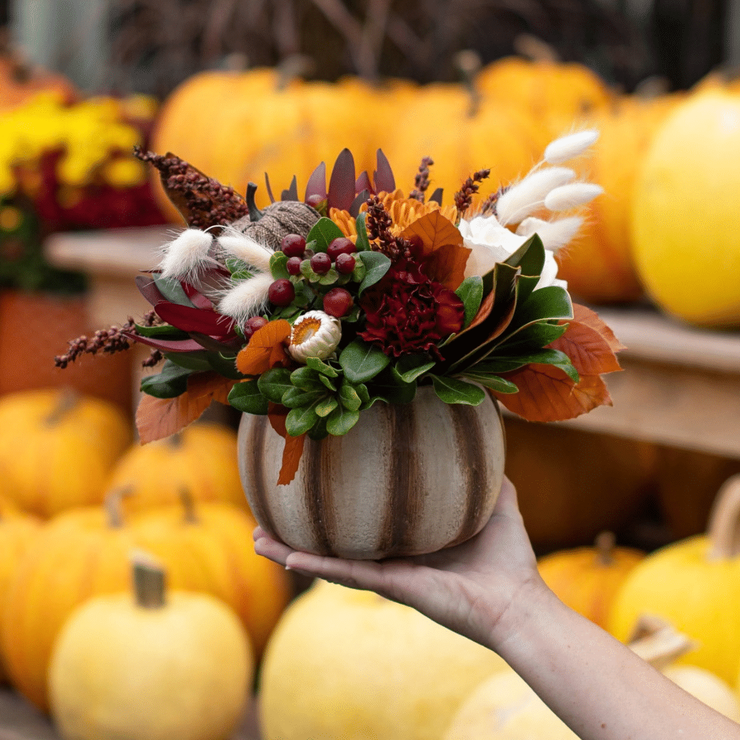 An outstretched hand holds a neutral brown, white, and burgundy fall floral arrangement in a cream and brown striped ceramic pumpkin.