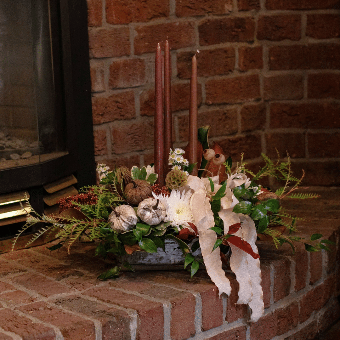 A fall floral arrangement with three brown taper candles, white mums, a cream-coloured ribbon bow, and other natural elements. It sits on a red brick fireplace.