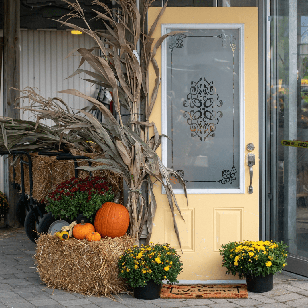 An image of a yellow door set up for fall with a corn stalk, straw bale, pumpkins, yellow mums, and a fall-themed welcome doormat.