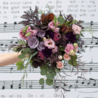 An outstretched hand holds a berry-toned bouquet with roses, carnations, lisianthus, snapdragons, etc. in front of a white and black background of music notes.