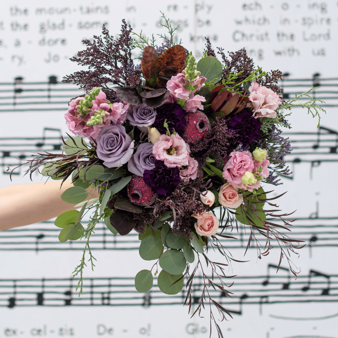 An outstretched hand holds a berry-toned bouquet with roses, carnations, lisianthus, snapdragons, etc. in front of a white and black background of music notes.