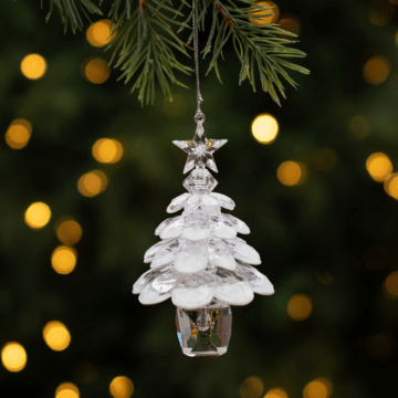 A clear and white acrylic ornament in the shape of a potted Christmas tree with a star on top hangs from an artificial branch with glowing warm white lights in the background.
