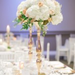 Elegant wedding table with tall floral centerpiece of white and blush flowers, gold accents, and a menu card.