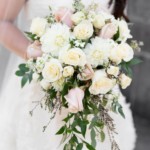 Bride holding elegant bouquet of white and blush roses with greenery, enhancing wedding attire's charm.