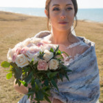Bride in lace dress holds bouquet of roses by a lake, wrapped in faux fur shawl on a sunny day.