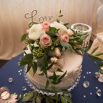 Elegant wedding cake with Love topper, adorned with pink roses and greenery on a crystal stand.
