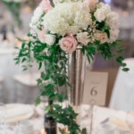 Elegant wedding table centerpiece with white and pink flowers in a tall vase, surrounded by table settings.